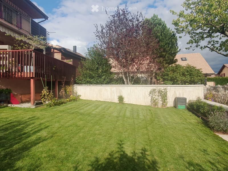Well-maintained lawn with trimmed grass, surrounded by trees, shrubs, and a low concrete wall. A wooden fence and a small shed are visible on the right side.