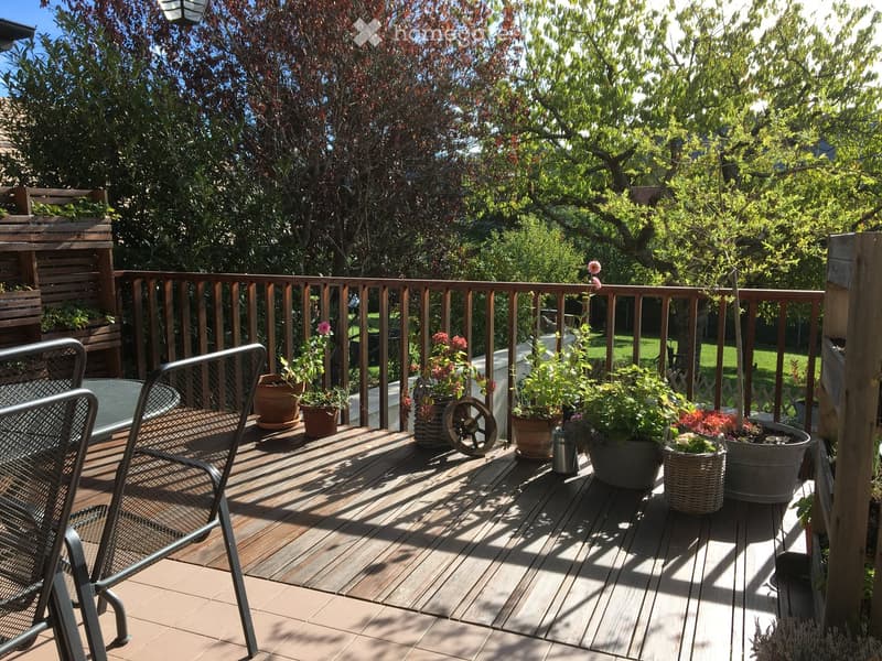 Wooden deck with railing, potted plants, and a metal chair, surrounded by trees and greenery.
