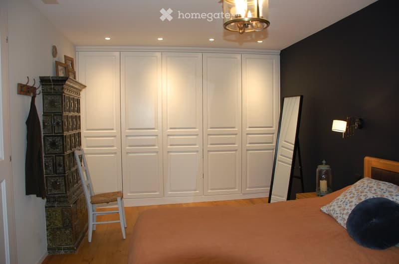A well-lit bedroom with a large bed, a white wardrobe, a wooden chair, a mirror, and a candle holder on a bedside table.