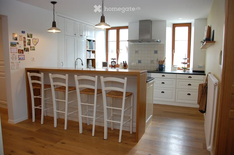 Bright, modern kitchen with white cabinets, wooden island, four bar stools, and various kitchen appliances. The space features a wooden floor, large windows, and a hanging pendant light.