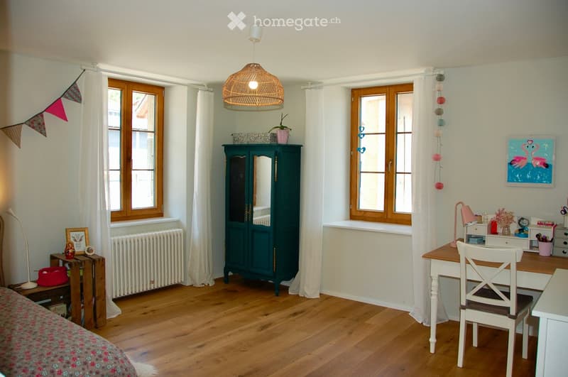 Cozy, well-lit room with wooden flooring, large windows, a bed, a wardrobe, a desk with a chair, and decorative items including a picture frame and a plant.
