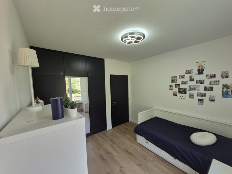 Modern bedroom with a single bed, white cabinet, floor lamp, potted plant, and decorative candle on a white dresser. Photos are displayed on the wall.
