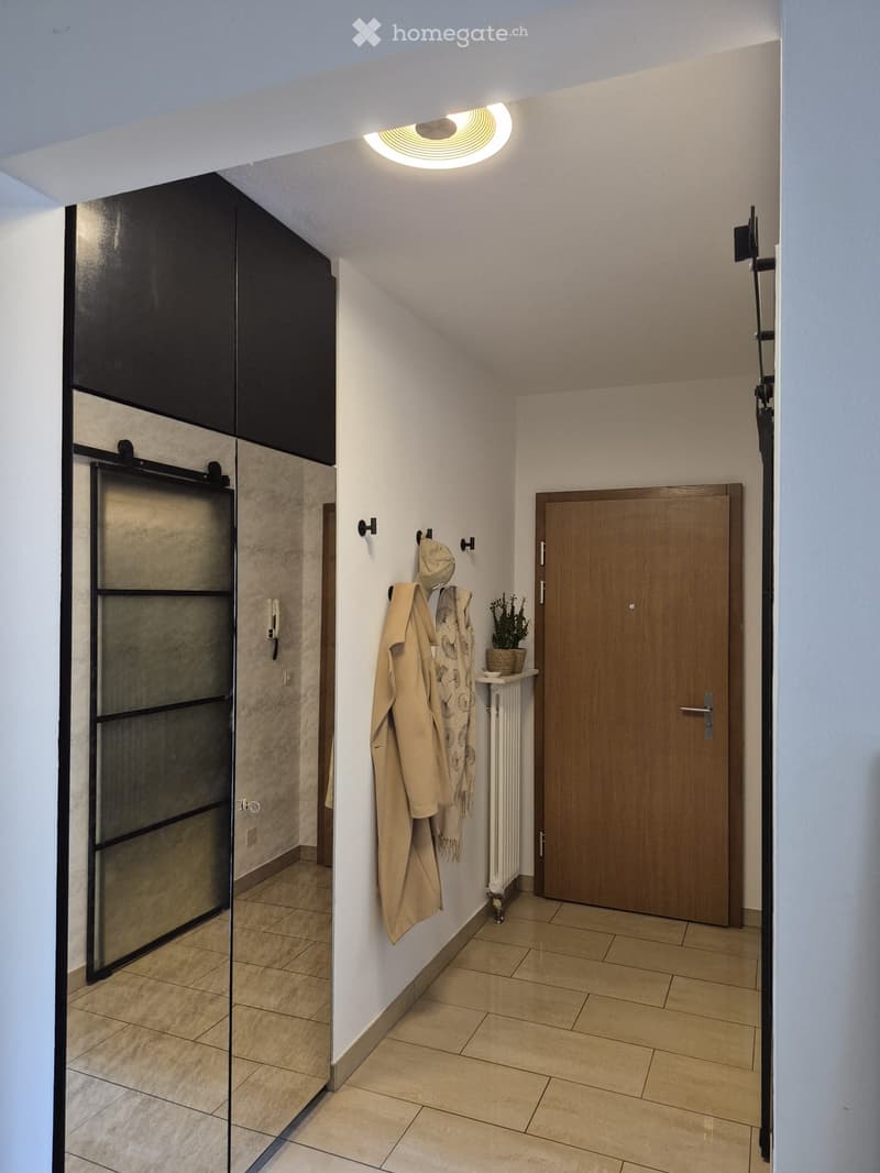 Modern hallway with tiled flooring, white walls, coat rack with coats and scarves, small potted plant, and a wooden door at the end.