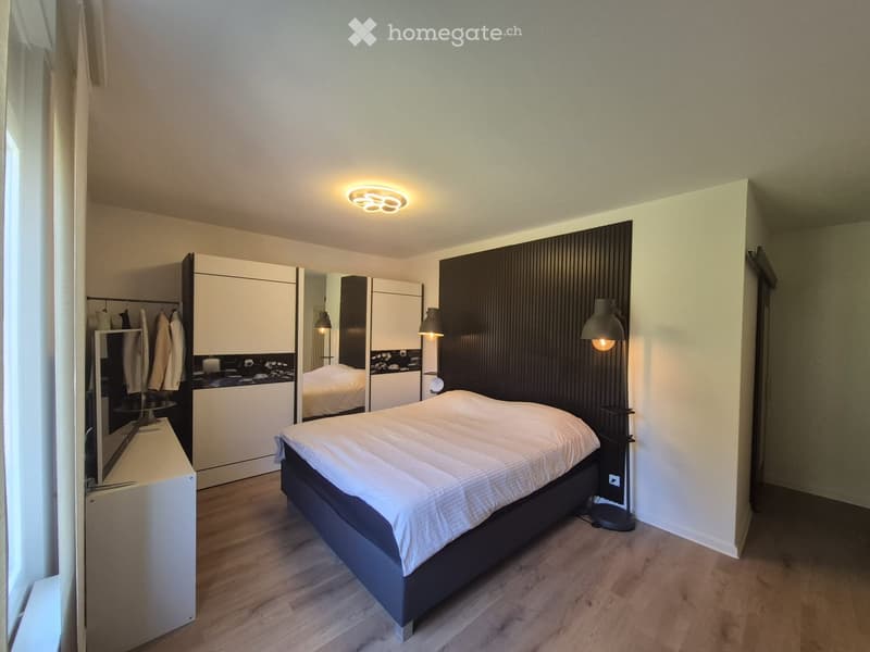 Modern bedroom with a large bed, wardrobe, mirror, and wall-mounted lamp. The room features wooden flooring and a sleek, contemporary design.