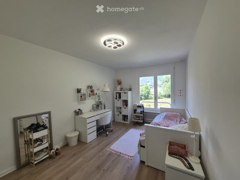 Cozy bedroom with a bed, desk, chair, mirror, shelves, and a nightstand. The room features wooden flooring, a large window with a view, and a round ceiling light.