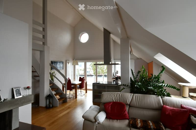 Bright, open living space with wooden flooring, a grey sofa with red cushions, a dining area with a table and chairs, a fireplace, and large windows offering a view of the outdoors.
