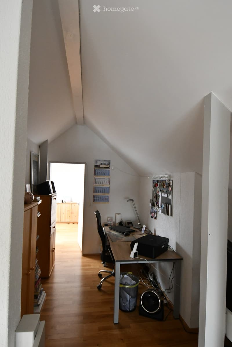 A small, well-lit attic room with wooden flooring, a desk with a laptop, a chair, a lamp, and a calendar on the wall. There is a cabinet and a fan visible.