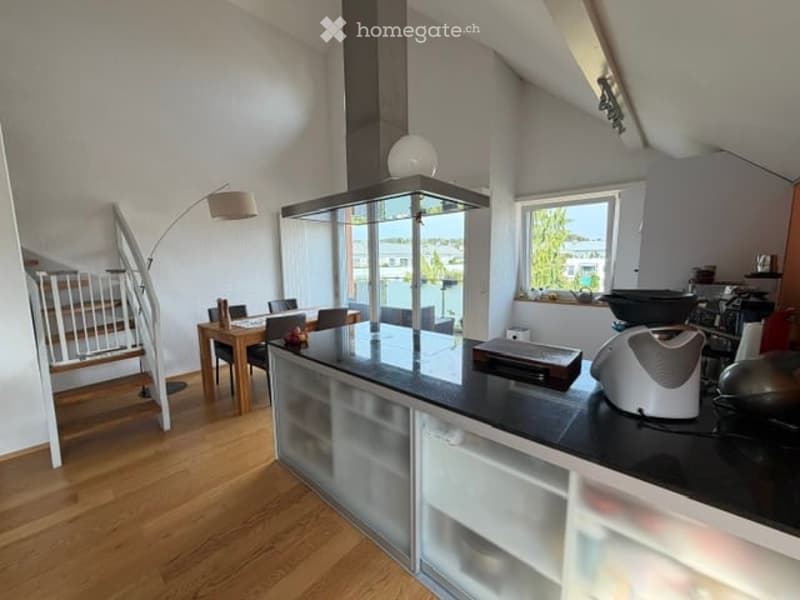 Modern kitchen with a central island, black countertops, and built-in cabinets. Includes a dining area with a wooden table and chairs, a floor lamp, and various appliances like a stove, microwave, toaster, and fridge. Large windows offer a view of the out