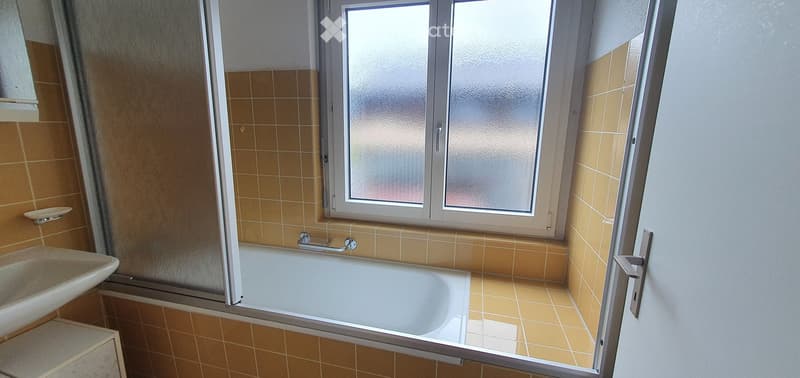 Bathroom with a bathtub, sink, and window with frosted glass. Beige tiled walls and floor.