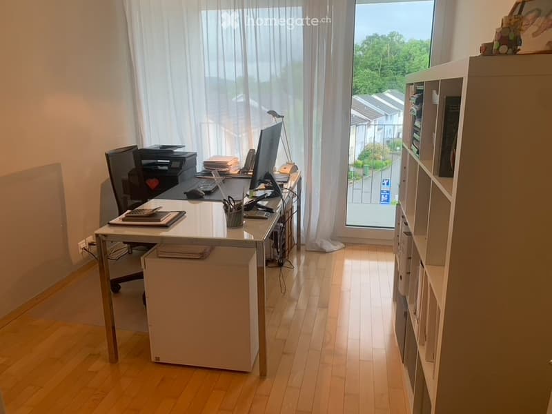 Desk area with computer monitor, office chair, shelf with books and a glass window with balcony