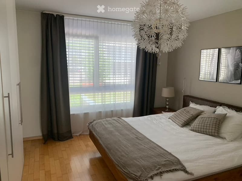 The bedroom is decorated with wooden flooring and a wooden bed, covered with a gray blanket and white sheets. The walls are cream-colored, and there are white window curtains and gray curtains over the window. A chandelier and a lamp are hanging from the