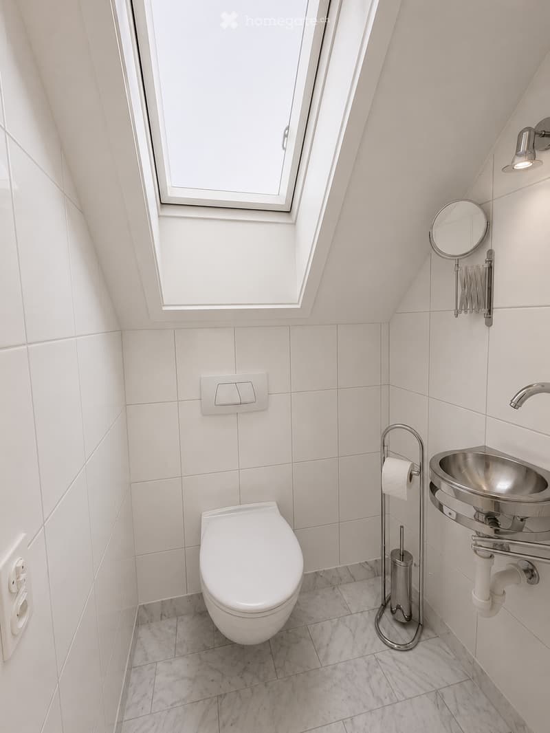 Modern bathroom with white tiled walls, marble flooring, a toilet, a stainless steel sink, a mirror, and a window with a skylight.