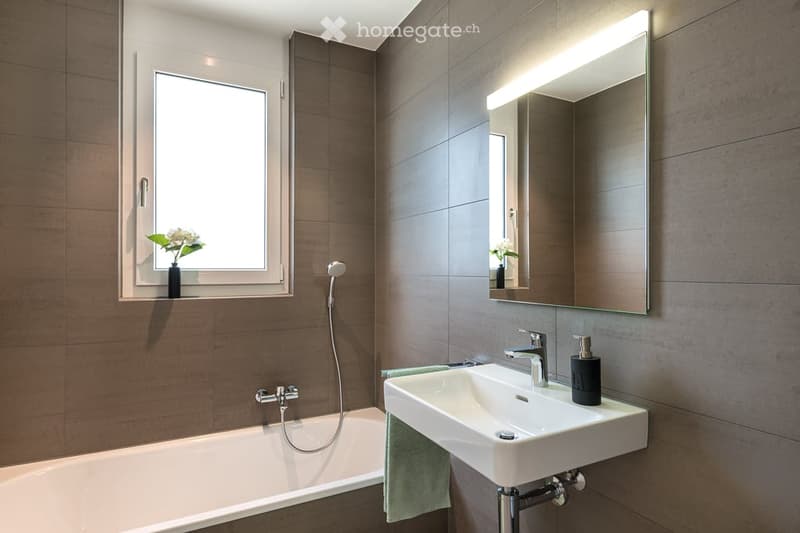 Modern bathroom with a bathtub, sink, mirror, and a small window with a vase of flowers. The walls are tiled in a dark brown color, and the lighting is bright.