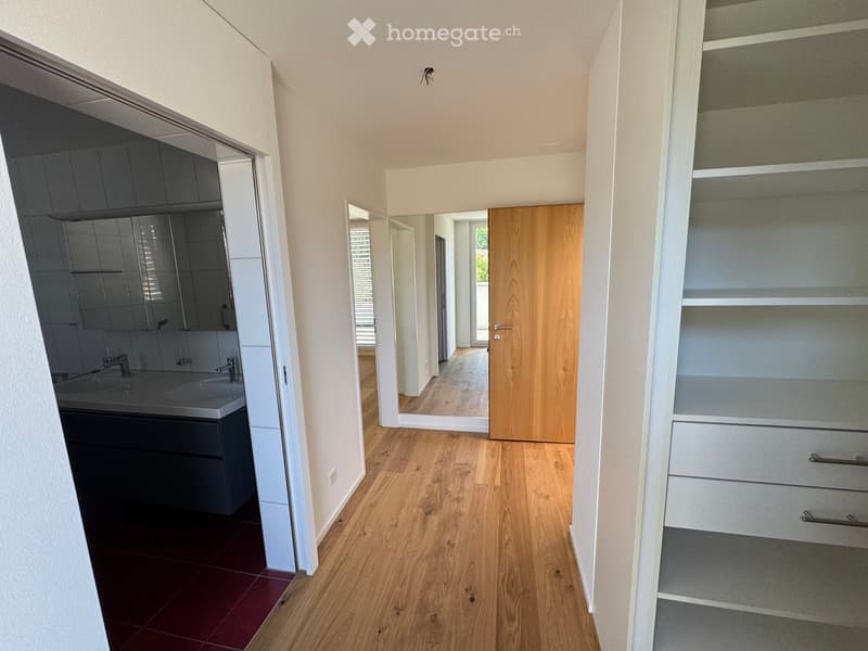 Empty hallway with wooden flooring, white walls, and a built-in closet on the right side. A bathroom is visible to the left with a sink, mirror, and dark cabinets.