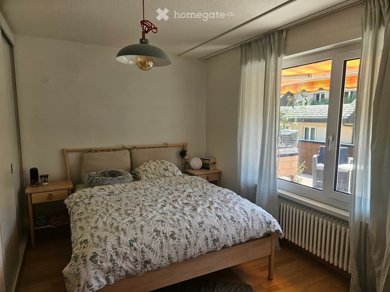 Cozy bedroom with wooden floor, large window with white curtains, bed with patterned duvet, nightstand, lamp, and small table with books and decorative items.