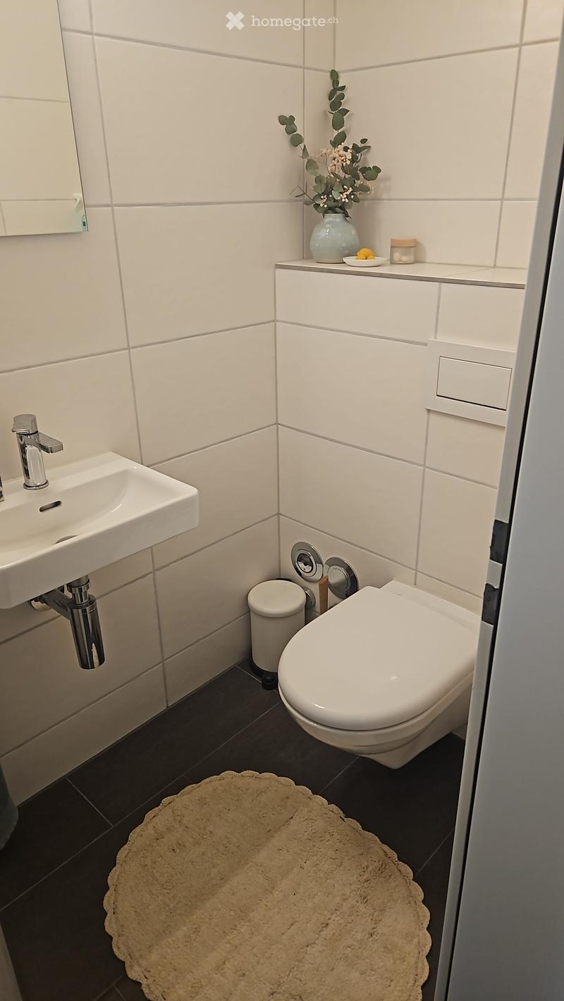 Modern bathroom with white tiles, a sink, toilet, and a small shelf holding a vase with flowers and a candle. A round, beige rug is placed in front of the toilet.