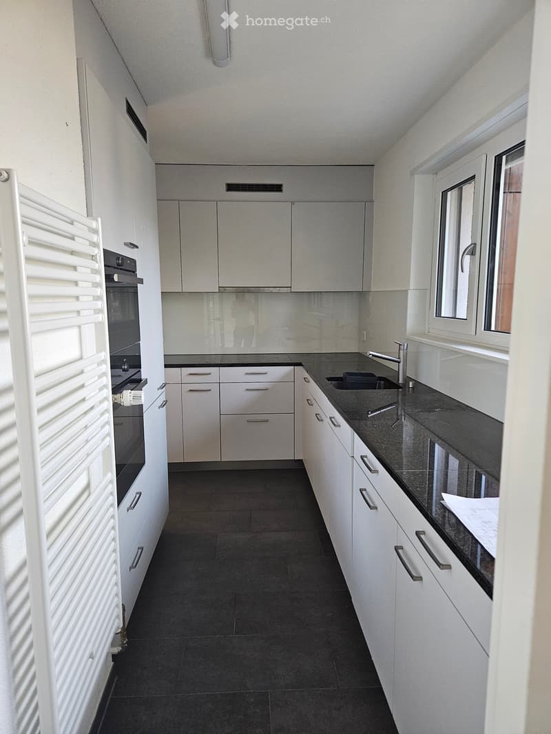 Modern kitchen with white cabinets, black countertops, and a stainless steel sink. Features include a built-in oven, a radiator, and a window with white frames.