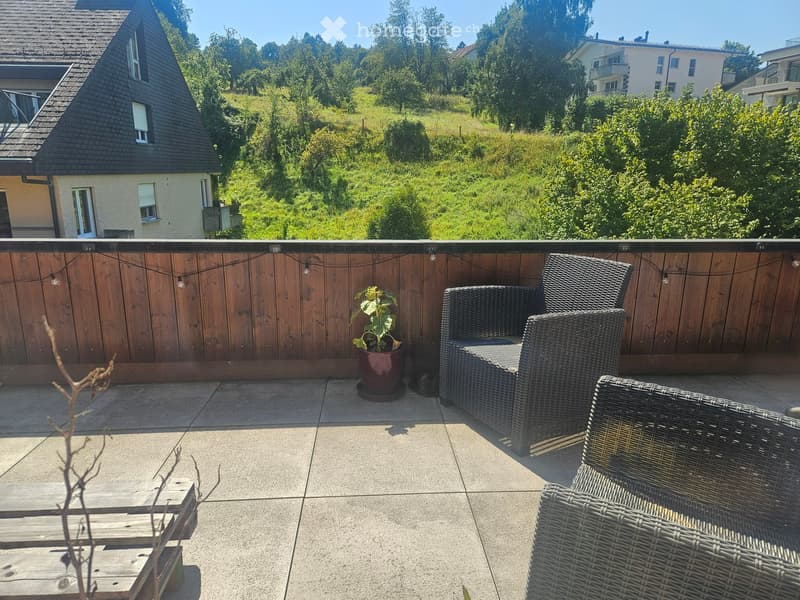 Balcony with wooden railing, wicker chairs, small table, potted plant, and scenic view of greenery and houses.