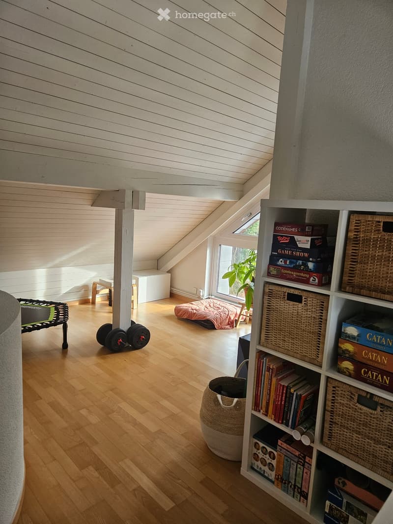 Cozy attic room with wooden floors, slanted white ceiling, large window, shelf with books and games, wicker basket, small bench, and weights.