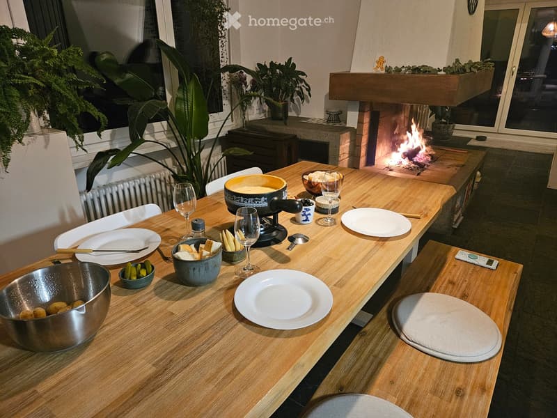 A cozy dining area with a long wooden table set for a meal, surrounded by chairs, a lit fireplace, and potted plants.