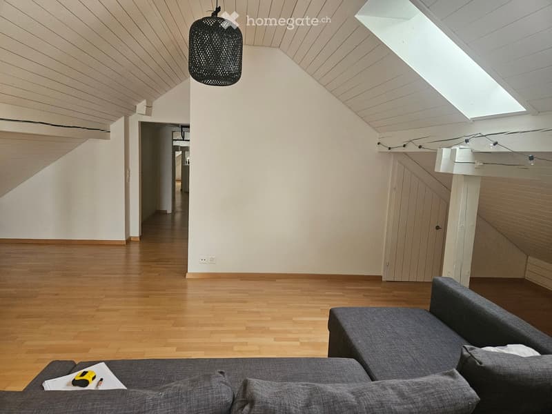 Modern living room with wooden flooring, white walls, and a grey sofa. A small table with a chair is visible, and a hanging lamp adorns the ceiling. The room features a skylight and a hallway leading to another area.