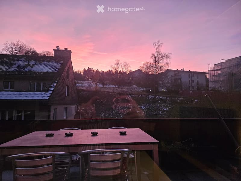 Balcony with a table and chairs, overlooking a snowy neighborhood at sunset.