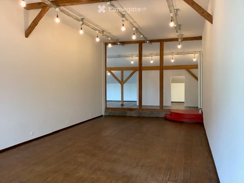 Empty room with wooden floor, white walls, exposed wooden beams, and track lighting. A red platform is visible in one corner.