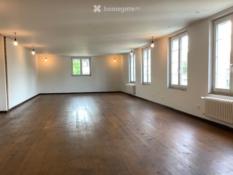 Open room, hardwood floors, white walls, large windows, hanging lamps, radiators.