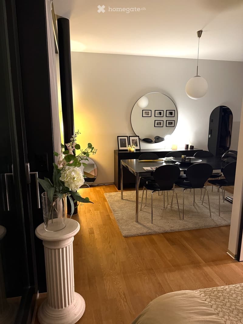 Modern dining area with wooden floor, round mirror, black chairs, glass table, and decorative vase on a white pedestal.