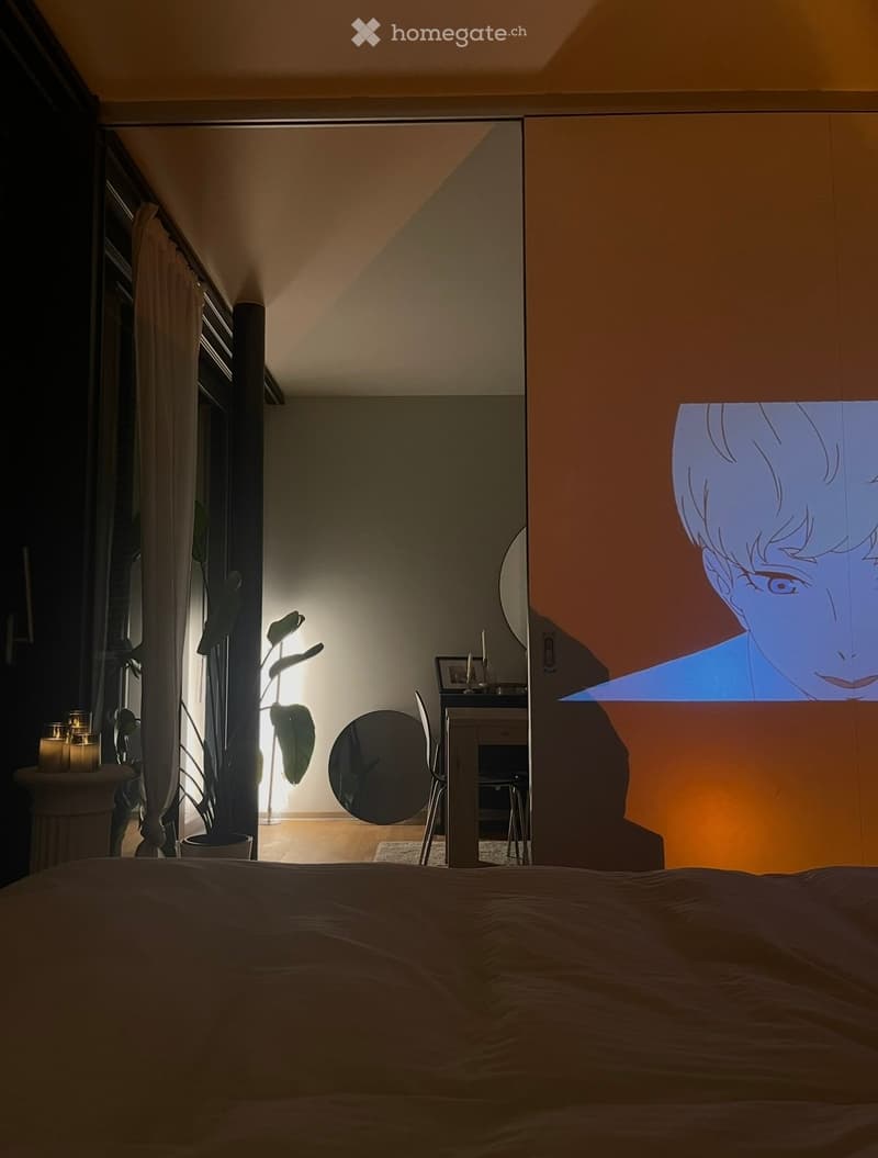 Bedroom with a large bed, a chair, a small table with a mirror, candles, and a potted plant. A projector screen displays an animated image on the wall.