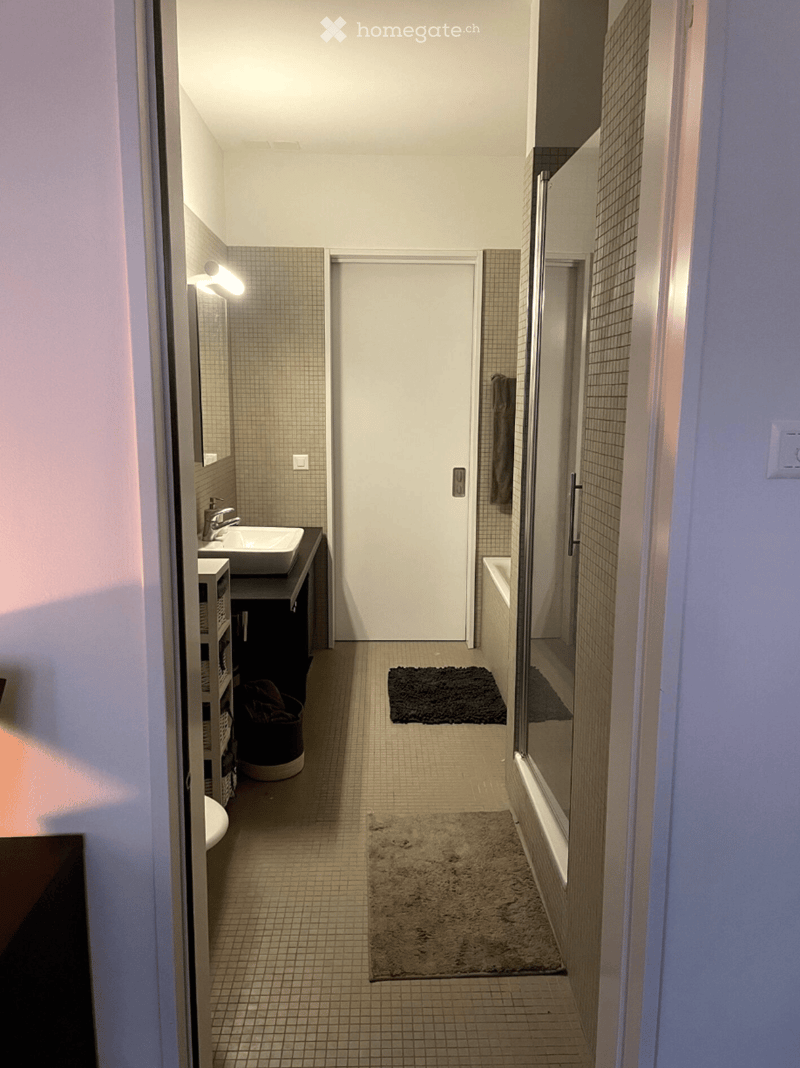 Modern bathroom with tiled walls and floor, a sink with a mirror, a bathtub, and two rugs placed on the floor.