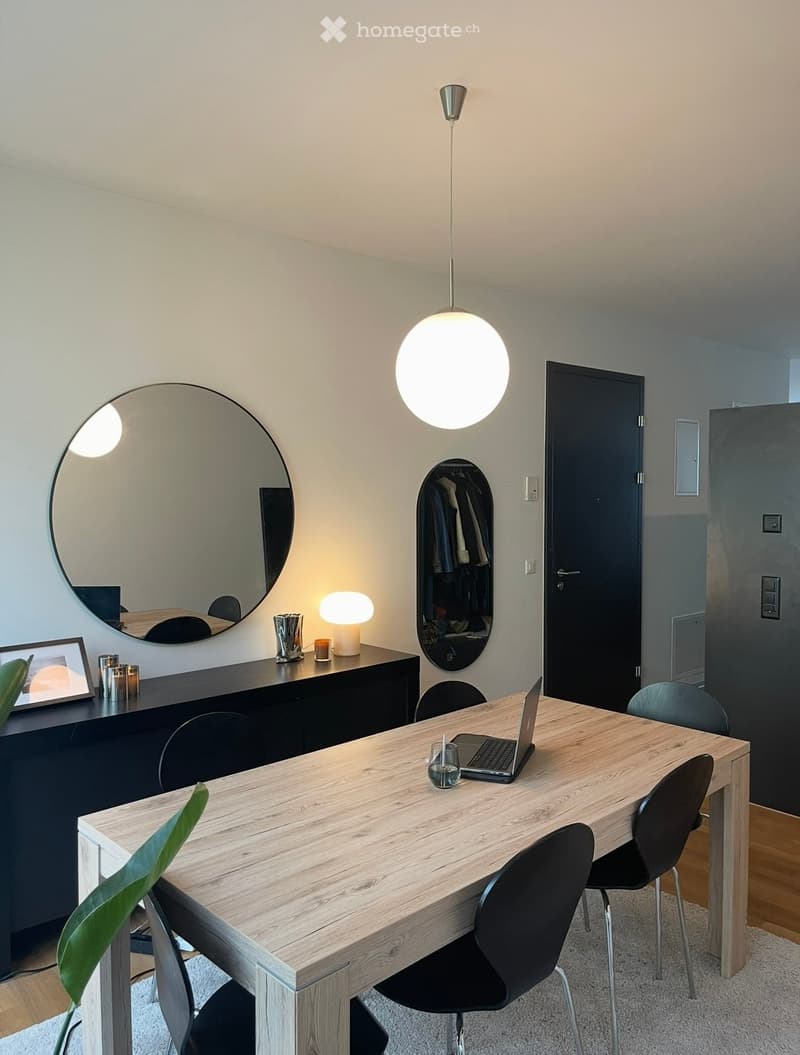 A modern, well-lit room with a wooden table, black chairs, a laptop, a large round mirror, a lamp, and a cabinet with decorative items.