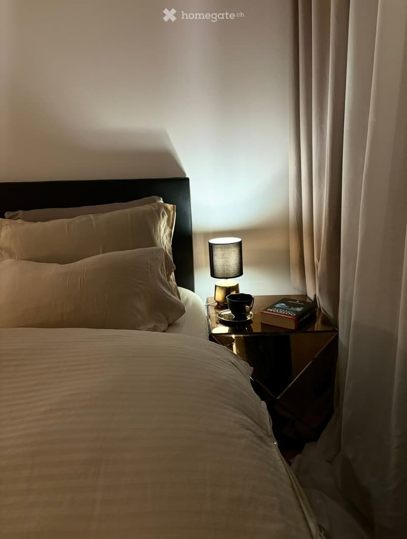 A neatly made bed with pillows and a duvet, a nightstand with a lamp, a book, and a cup, and a window with curtains letting in soft light.