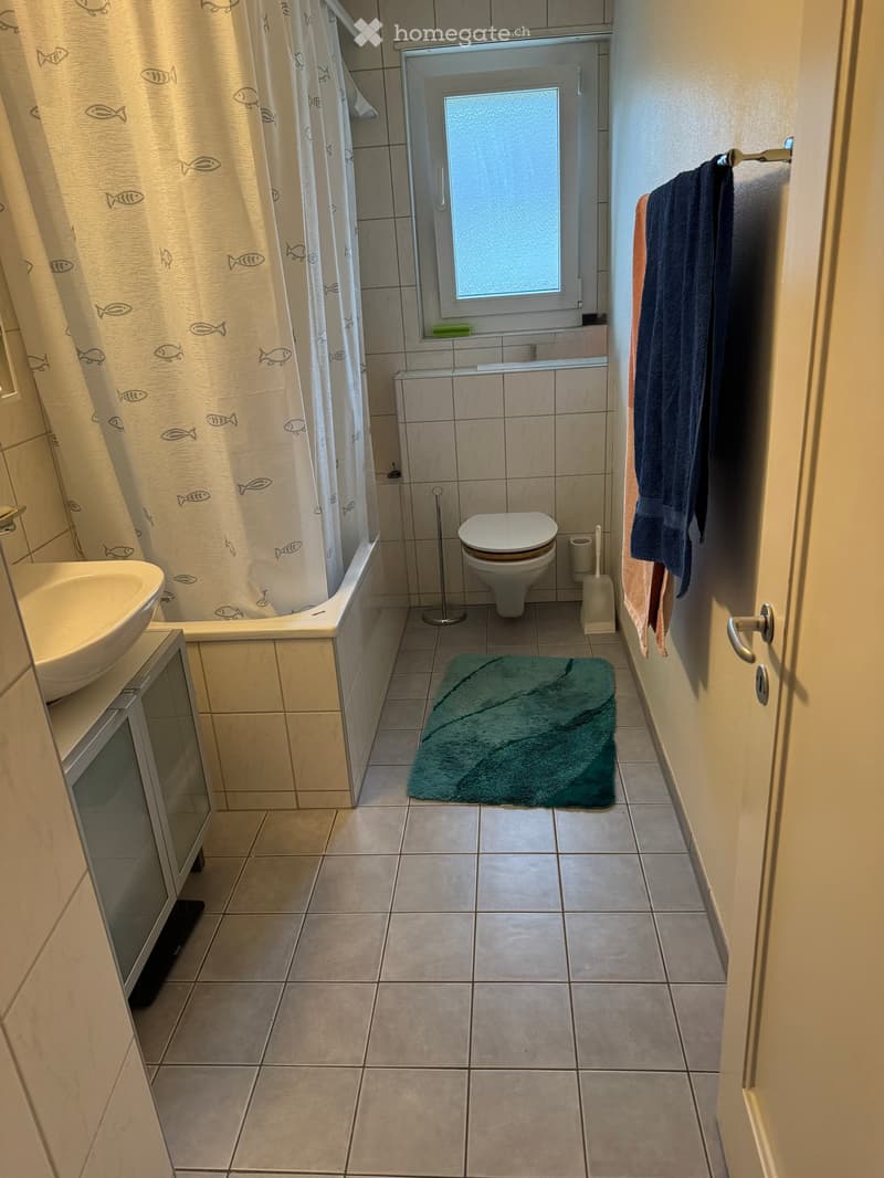 Bathroom with tiled walls and floor, bathtub with fish-patterned shower curtain, toilet, sink, mirror, and towels on a rack.