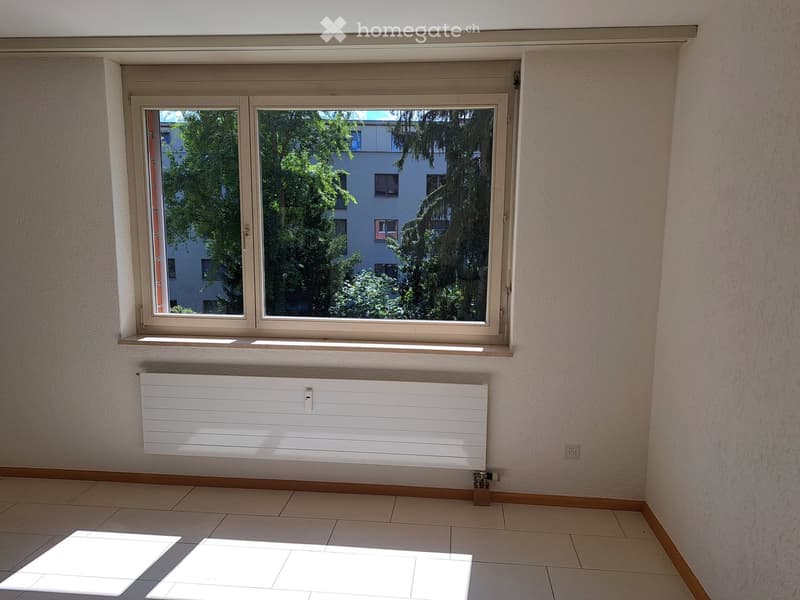 Empty room with tiled floor, white radiator, and large window with view of trees and buildings.