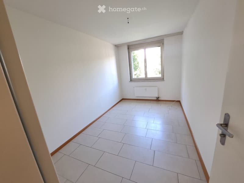 Empty room with white tiled floor, white walls, and a window with a view of greenery. The room has a simple wooden baseboard and a door with a silver handle visible on the right side.