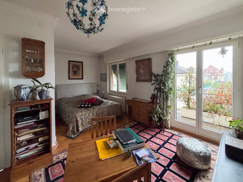 A cozy living space with a bed, a wooden table, a bookshelf, and a large window leading to a balcony. The room features a patterned rug, a chandelier, and several potted plants.