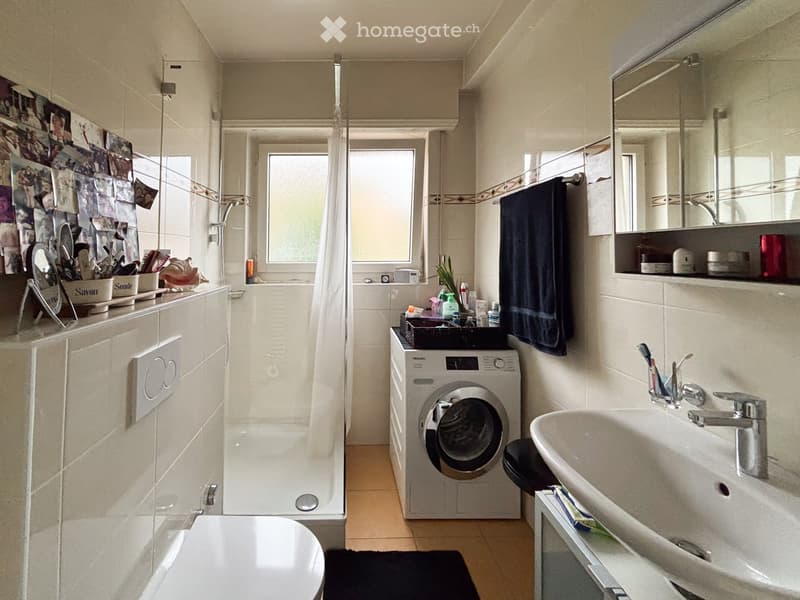 Modern bathroom with a toilet, sink, mirror, shower, and washing machine. Features a towel, toothbrush holder, and various toiletries on the sink counter.
