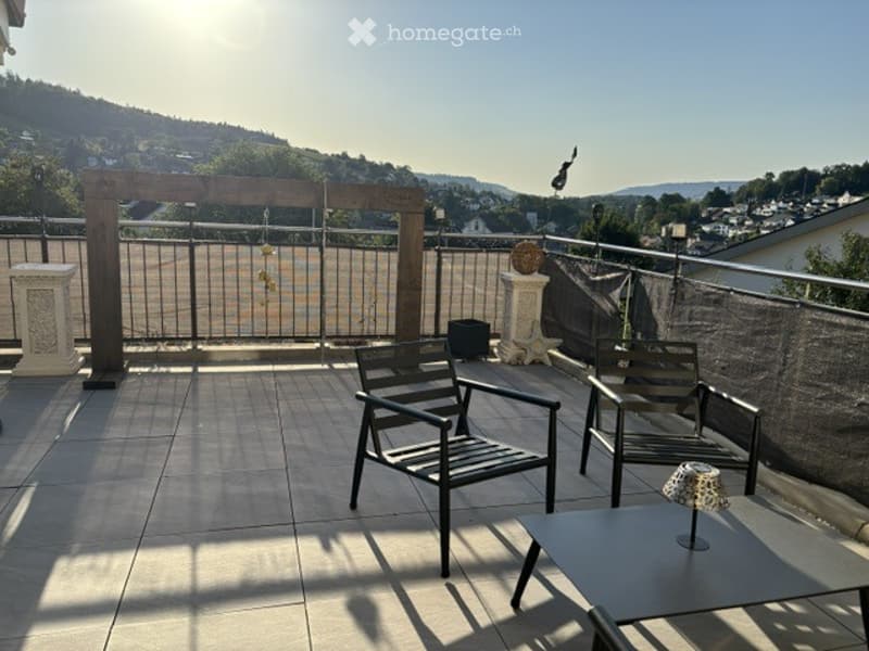 Balcony with tiled floor, metal railing, two chairs, small table, and scenic view of a town and hills.