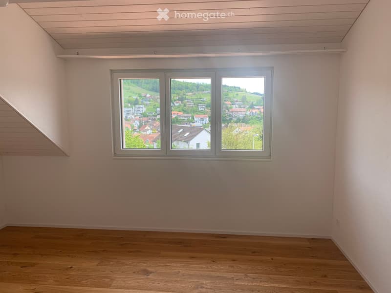 Empty room with wooden flooring, white walls, and large windows offering a view of a town and green landscape.