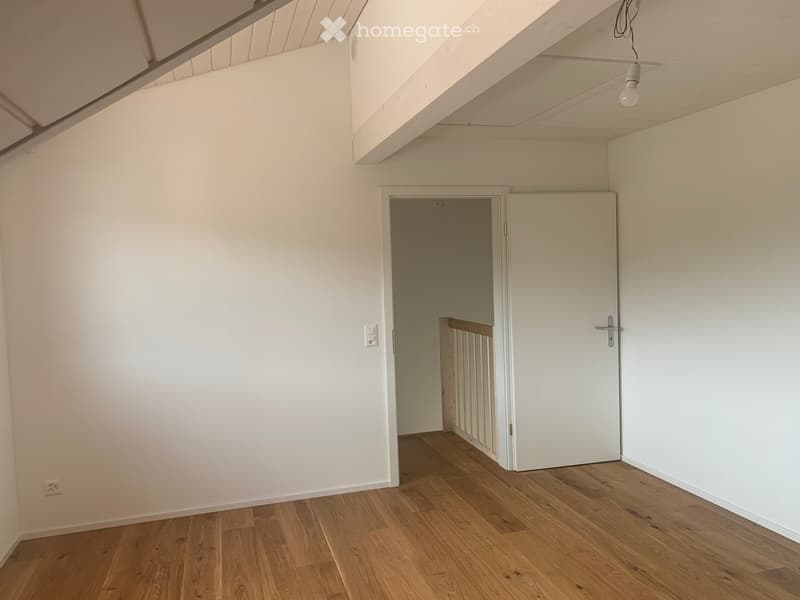 Empty room with wooden flooring, white walls, and a doorway leading to another room. Exposed wooden beams on the ceiling.