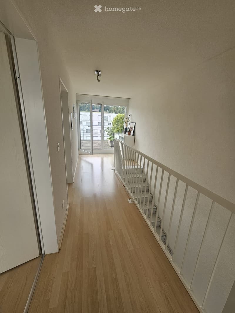 A well-lit hallway with wooden flooring, white walls, and a staircase leading upwards. A small table with a plant and picture frame is visible near the open sliding glass door at the end of the hallway.