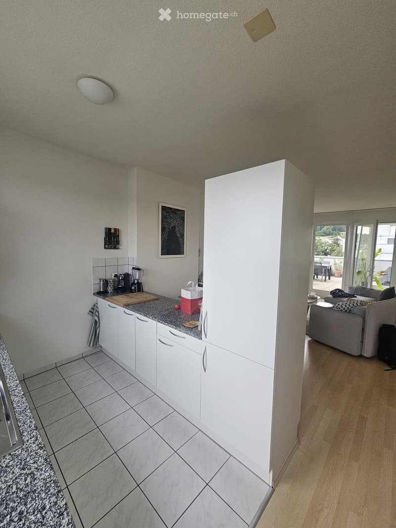 A modern kitchen with white cabinets, a granite countertop, and various appliances, connected to a living area with a sofa and a small table.