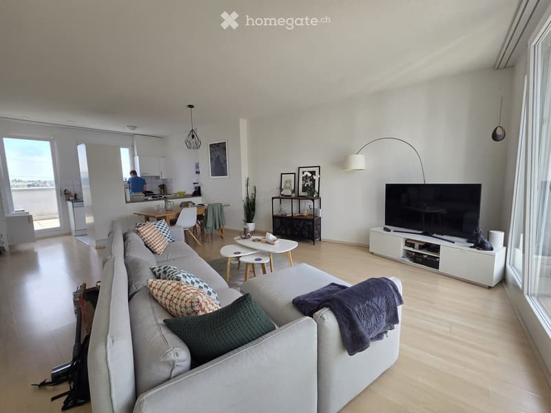Modern living room with a grey sectional sofa, coffee table, TV stand, and a small dining area with a table and chairs. The room features large windows, wooden flooring, and contemporary decor including a floor lamp and picture frames.
