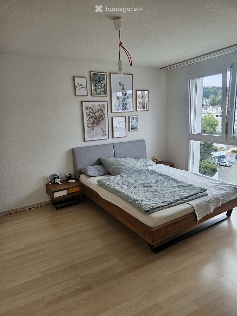 Modern bedroom with wooden flooring, a large bed with grey bedding, a nightstand, and several picture frames on the wall. A window offers a view of the outside.