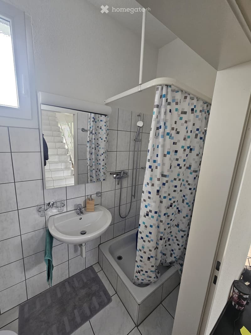 Modern bathroom with a white sink, bathtub, and shower curtain. Features a large mirror, tiled walls, and a small window.