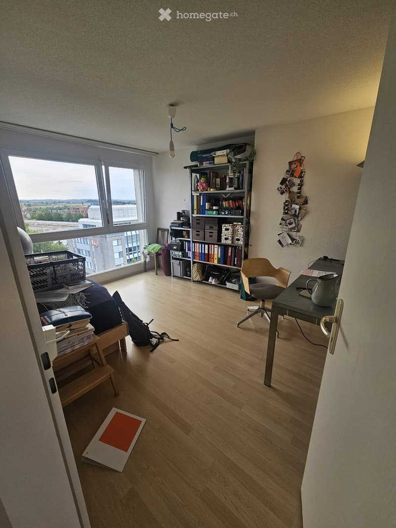 A furnished room with wooden flooring, large windows offering a view, a sofa, a small wooden stool, a chair, a table with a teapot, and a bookshelf filled with books and storage boxes.