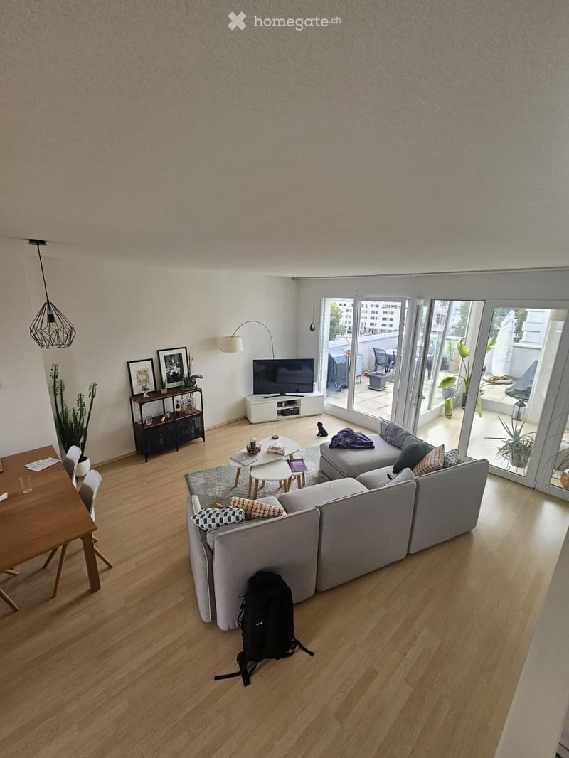 Modern living room with light wooden flooring, grey sectional sofa, coffee table, dining table, TV, and large sliding glass doors leading to a balcony.