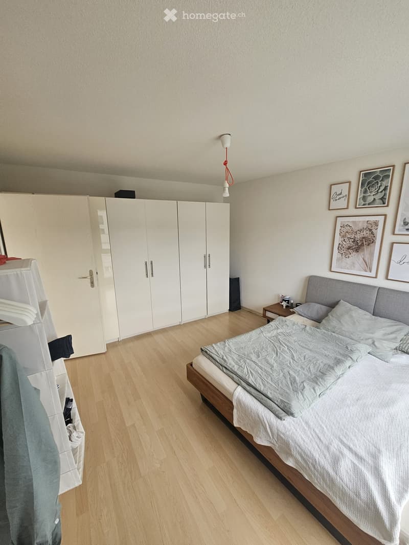 Modern bedroom with a wooden floor, a large bed with grey bedding, a white wardrobe, a nightstand, and several framed pictures on the wall.