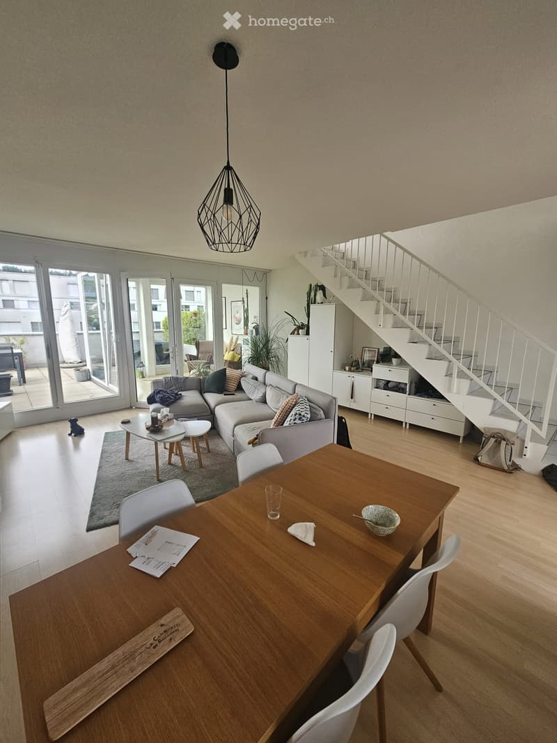 Open-plan living and dining area with a grey sectional sofa, coffee table, dining table with chairs, pendant light, and a staircase leading to an upper level.